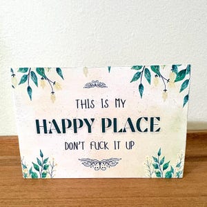 May include: A rectangular sign with the text "THIS IS MY HAPPY PLACE DON'T FUCK IT UP" in a bold, dark teal font. The sign has a cream-colored background with green and yellow floral accents.