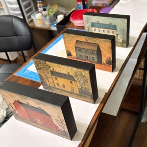 Four primitive Americana decor blocks displayed together on a white shelf in a craft room, featuring vintage colonial saltbox homes in distressed blue, red, cream, and mustard tones with early American farmhouse style.