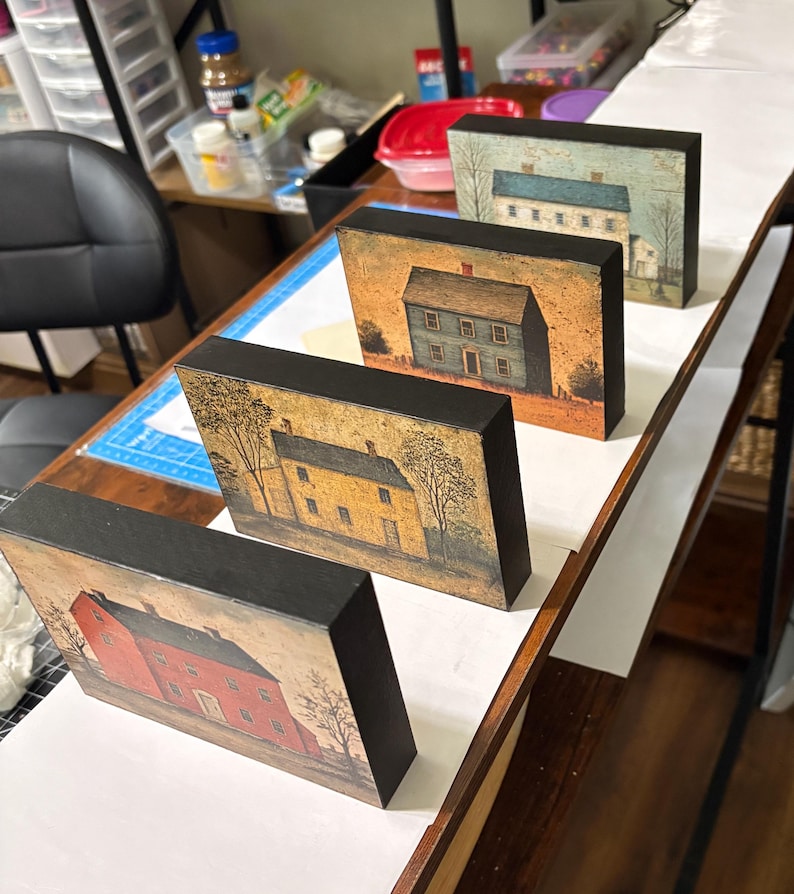 Four primitive Americana decor blocks displayed together on a white shelf in a craft room, featuring vintage colonial saltbox homes in distressed blue, red, cream, and mustard tones with early American farmhouse style.