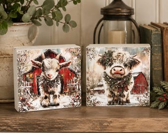 May include: Two square wooden blocks featuring printed artwork. The left block shows a young goat wearing a wreath in front of a red barn. The right block displays a fluffy Highland cow wearing a wreath in front of a red barn in a snowy setting.