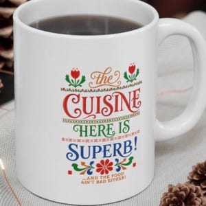May include: A white ceramic mug featuring a colorful graphic design with red, green, and blue text that reads: the CUISINE HERE IS SUPERB! ...AND THE FOOD AIN'T BAD EITHER! The design includes decorative floral elements and geometric patterns.