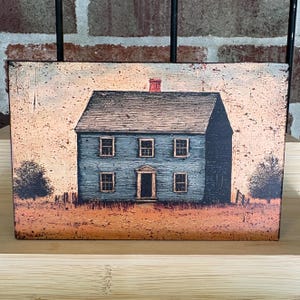 Primitive Americana decor block featuring a distressed blue colonial saltbox house with black roof and rustic countryside background, displayed on a wooden shelf against a brick wall.