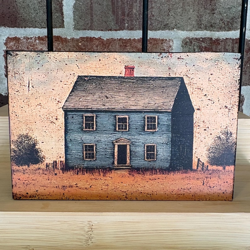 Primitive Americana decor block featuring a distressed blue colonial saltbox house with black roof and rustic countryside background, displayed on a wooden shelf against a brick wall.