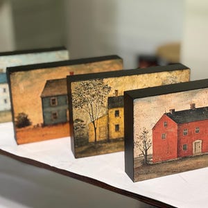 Four primitive Americana decor blocks displayed together on a white shelf in a craft room, featuring vintage colonial saltbox homes in distressed blue, red, cream, and mustard tones with early American farmhouse style.