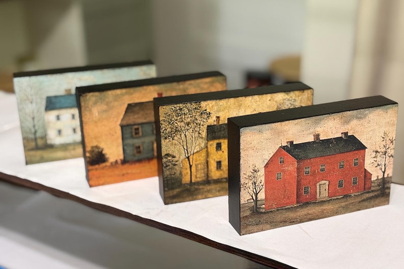 Four primitive Americana decor blocks displayed together on a white shelf in a craft room, featuring vintage colonial saltbox homes in distressed blue, red, cream, and mustard tones with early American farmhouse style.