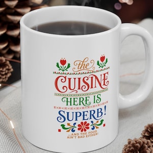 May include: A white ceramic mug featuring colorful text that reads: the CUISINE HERE IS SUPERB! ...AND THE FOOD AIN'T BAD EITHER! The design includes red, green, and blue lettering with decorative floral and geometric patterns in a folk-art style.