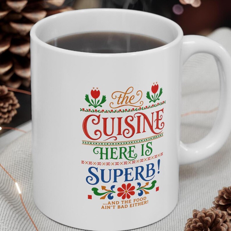 May include: A white ceramic mug featuring colorful text that reads: the CUISINE HERE IS SUPERB! ...AND THE FOOD AIN'T BAD EITHER! The design includes red, green, and blue lettering with decorative floral and geometric patterns in a folk-art style.