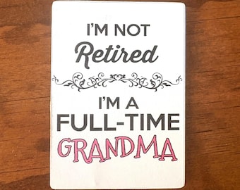 May include: A rectangular white sign with black and pink text that reads I'M NOT Retired I'M A FULL-TIME GRANDMA. The sign features a decorative black flourish element between the two lines of text, centered on a flat, smooth surface.