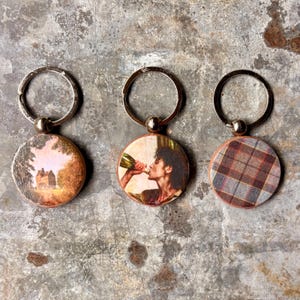May include: Three round keychains with silver rings. One features a landscape painting of a building, another depicts a person drinking from a bottle, and the last has a red, brown, and gray plaid pattern.