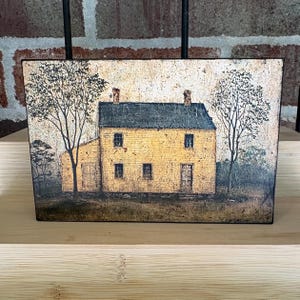 Primitive country decor block with mustard yellow colonial farmhouse artwork, distressed vintage finish, and rustic Americana style displayed on a wooden shelf against a brick wall.