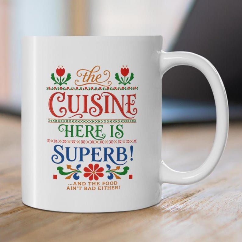 May include: A white ceramic mug featuring colorful decorative text that reads: the CUISINE HERE IS SUPERB! ...AND THE FOOD AIN'T BAD EITHER! The design includes red, green, and blue floral motifs and geometric patterns arranged in a vertical layout.