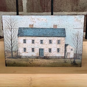 Early American farmhouse decor block featuring a distressed cream colonial saltbox home with black roof and rustic primitive landscape, displayed on a wooden shelf.