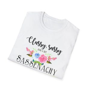 May include: White t-shirt with the text "Classy, Sassy, and a bit Sassenachy" in black script. The shirt features a floral design with pink, blue, and white flowers. The t-shirt is folded and displayed on a white background.