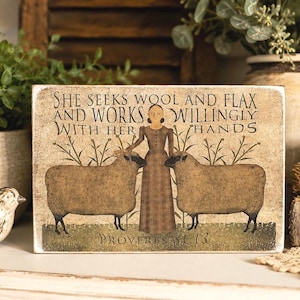 May include: A rectangular wooden sign with a distressed finish featuring an illustration of a person in a brown dress standing between two brown sheep. The text reads: SHE SEEKS WOOL AND FLAX AND WORKS WILLINGLY WITH HER HANDS PROVERBS 31:13.