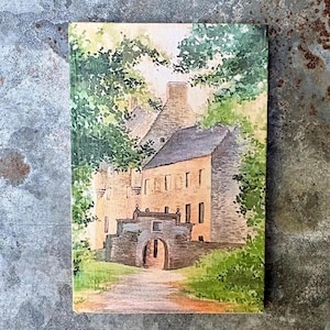 May include: A rectangular wooden plaque featuring a watercolor painting of a stone building with a prominent arched gateway. The scene includes lush green trees surrounding the structure, with a light-colored path leading toward the entrance.