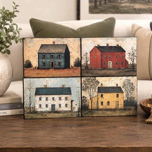 Four primitive Americana decor blocks displayed together on a wooden table in an elegant living room, featuring vintage colonial saltbox homes in distressed blue, red, cream, and mustard tones with early American farmhouse style.