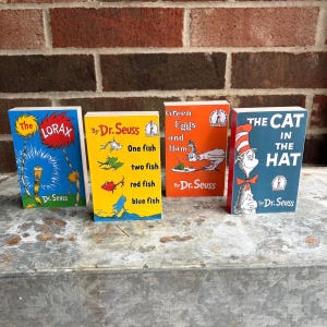 May include: Four Dr. Seuss books standing upright. The blue book is titled The Lorax. The yellow book is titled One fish two fish red fish blue fish. The orange book is titled Green Eggs and Ham. The blue book is titled The Cat in the Hat.