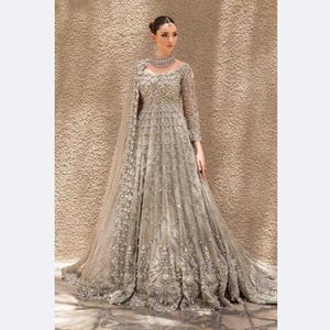 Puede incluir: Un vestido largo plateado con intrincados bordados y abalorios. El vestido presenta mangas largas, un cuello alto y una falda completa que se arrastra por detrás. Un chal a juego está drapeado sobre los hombros.