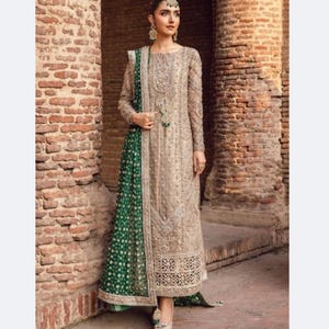 Könnte beinhalten: Ein langes, beigefarbenes formelles Kleid mit aufwendiger Stickerei und Verzierungen. Das Kleid hat lange Ärmel und ein detailliertes Oberteil. Ein grün-goldenes Schal mit Muster ist über einer Schulter drapiert. Das Kleid wird mit silbernen Absätzen kombiniert.