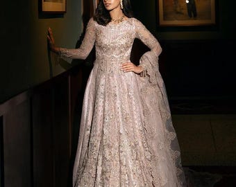 Puede incluir: Un vestido formal largo de color rosa claro con intrincados bordados y mangas largas. El vestido presenta una falda fluida y una delicada superposición de encaje. La modelo lleva joyas de oro. El vestido es adecuado para ocasiones especiales.