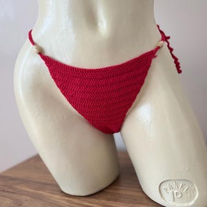 May include: A red crochet bikini bottom with wooden bead accents. The handmade swimwear features a classic triangle cut and adjustable ties. The vibrant red color and textured stitch create a stylish beachwear piece.