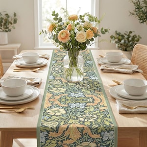 May include: A dining table set for a meal, featuring a floral patterned table runner in shades of green, blue, and gold. The table is set with white plates, bowls, gold cutlery, and linen napkins. A vase of flowers sits in the center.