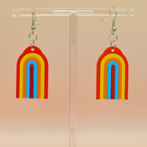 May include: A pair of colorful rainbow earrings. Each earring features a red arch with layers of yellow, orange, blue, and yellow. The earrings hang from silver-colored fish hook ear wires. The earrings are displayed on a clear plastic stand.