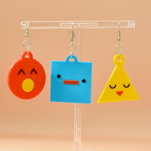 May include: Three colorful, cartoon-style earrings on display. The earrings are a red circle with a yellow circle, a blue square, and a yellow triangle. Each earring has a face design and is suspended from a clear display stand.
