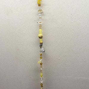 Puede incluir: Una larga cadena de cuentas con cuentas transparentes, amarillas y doradas. La cadena tiene un cristal transparente en la parte inferior y un dije de flor plateada.