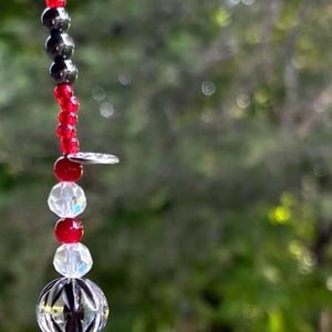 Puede incluir: Adorno de cuentas con cuentas rojas, negras, transparentes y marrones. El adorno tiene una mezcla de formas y texturas, incluyendo cuentas redondas, cuentas facetadas y un elemento decorativo plateado. El fondo es verde borroso.