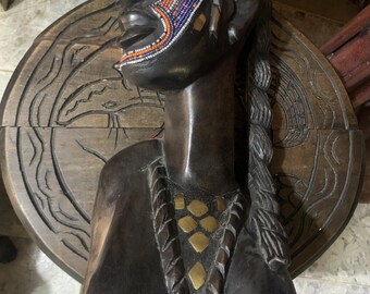 May include: A dark wood bust of a woman with a beaded face and a gold necklace. The bust is carved with intricate details and is a beautiful example of African art.