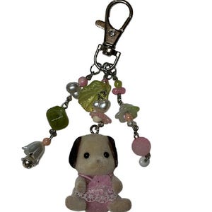 May include: A keychain featuring a small flocked dog figurine wearing a pink outfit with lace trim. The dog hangs from a silver-toned lobster clasp attached to three beaded chains adorned with green leaf-shaped beads, pink spheres, and white bell charms.