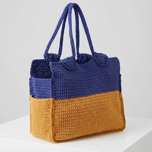 Può includere: Borsa tote all'uncinetto fatta a mano in un design bicolore. La metà superiore è blu scuro e la metà inferiore è giallo dorato. La borsa ha due robusti manici blu. La borsa è su una superficie bianca.