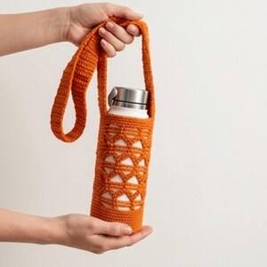 May include: An orange crochet water bottle holder with a strap. The holder has a geometric design and is holding a white stainless steel water bottle with a silver lid. The holder is being held up by a person's hands.