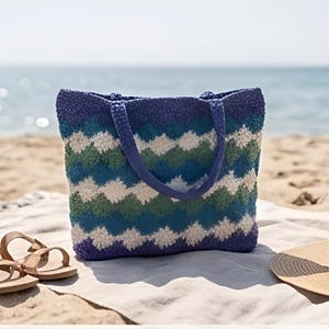 May include: A blue, green, and white patterned tote bag with matching handles sits on a beach towel. Brown sandals and a straw hat are next to the bag. The ocean is visible in the background.