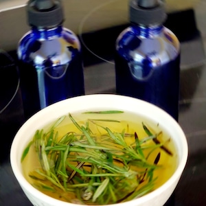 May include: Two cobalt blue glass bottles with black droppers are positioned behind a white bowl filled with rosemary sprigs in a yellow liquid. The bowl is white with a simple design. The items are on a dark surface.