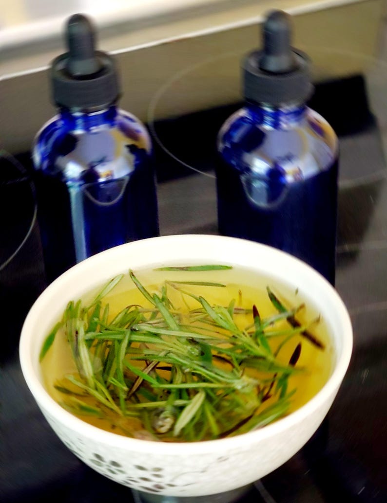 May include: Two cobalt blue glass bottles with black droppers are positioned behind a white bowl filled with rosemary sprigs in a yellow liquid. The bowl is white with a simple design. The items are on a dark surface.