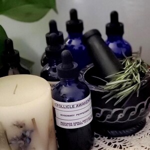 May include: A collection of apothecary items, including several dark blue glass bottles with droppers, a black mortar and pestle, a white candle, and sprigs of rosemary. The bottles have labels that read "Follicle Awakening".