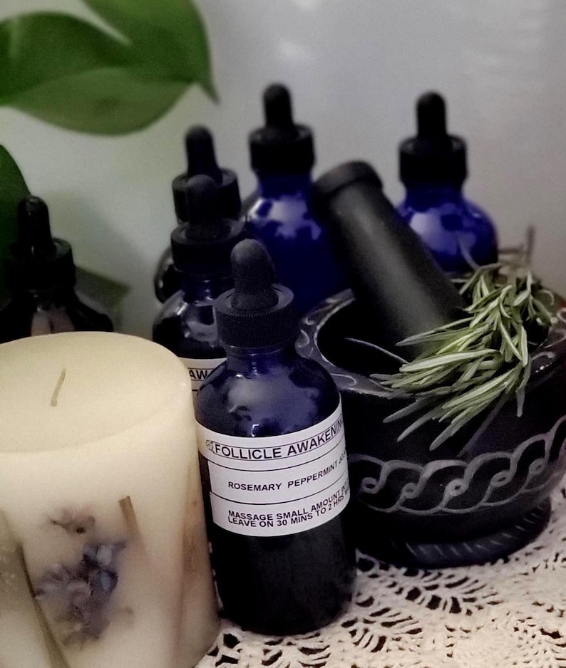May include: A collection of apothecary items, including several dark blue glass bottles with droppers, a black mortar and pestle, a white candle, and sprigs of rosemary. The bottles have labels that read "Follicle Awakening".