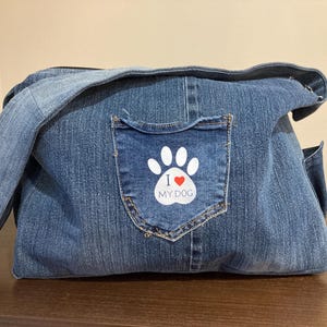May include: A blue denim dog carrier bag with a shoulder strap. The bag features a pocket with a white paw print design and the text "I ❤️ MY DOG". The bag is made from recycled denim.