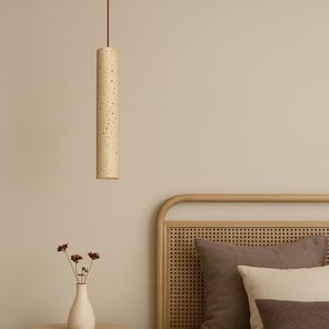 Puede incluir: Una lámpara colgante cilíndrica beige con un patrón moteado cuelga de un cable marrón. La luz está sobre una escena de dormitorio con una mesa auxiliar redonda, un jarrón con flores secas y una cama con almohadas.