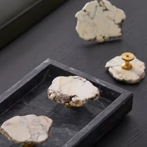May include: Cabinet knobs made from natural stone, featuring brass fittings. The knobs have an irregular shape and display a mix of white, cream, and brown colours. They are arranged on a dark grey surface, with some placed in a rectangular tray.