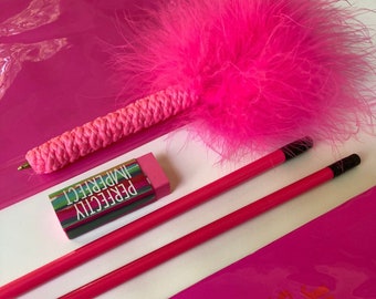 Op de afbeelding: Een roze verenpen, twee roze potloden en een roze gum met de tekst "PERFECTLY IMPERFECT" erop gedrukt. De items liggen op een roze oppervlak.