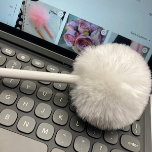 Op de afbeelding: Een witte, pluche pompon sleutelhanger bevestigd aan een witte pen. De pen ligt op een grijs toetsenbord met zwarte toetsen. Het toetsenbord maakt deel uit van een tabletcomputer.