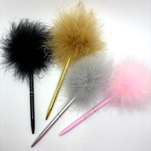 May include: Four pens with fluffy feather toppers in black, gold, silver, and pink. The pens have sleek, metallic barrels. The feather toppers are round and voluminous, adding a touch of whimsy. The pens are arranged on a white surface.