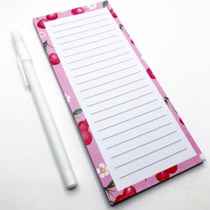 May include: A pink notepad with a cherry and flower pattern on the cover. The notepad has white lined paper and comes with a white pen.