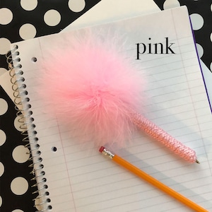 Op de afbeelding: Een roze pluche pen met een gevlochten koord en het woord "pink" geschreven op een notitieboek.