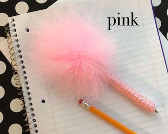 Op de afbeelding: Een roze pen met een grote, pluizige veren pompon en een textuurrijke, gehaakte roze garenwikkel rond de houder. Het woord pink staat in zwarte letters boven de pen, die op een gelinieerd notitieblok naast een geel potlood ligt.