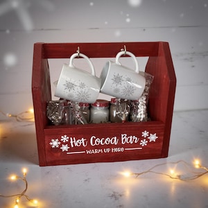 May include: A red wooden hot cocoa bar caddy with two white mugs decorated with snowflakes hanging from hooks. The caddy holds small jars and bags of cocoa ingredients. The words "Hot Cocoa Bar - Warm Up Here" are printed on the front.