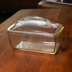 Puede incluir: Una fuente de horno rectangular de vidrio transparente con asa en la tapa, sobre una mesa de madera marrón oscuro. El plato tiene un borde teñido de amarillo. El fondo muestra un salón con muebles y decoración.